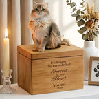 A fluffy cat sits on a wooden PawPrint Cat Urn engraved with "No longer by my side but Forever in my Heart, Momo, 2026," next to a ceramic cat memorial, a lit candle, dried flowers, and a framed photo on the table.