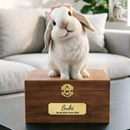 Bunny Sweet Memories is a lifelike 3D printed rabbit statue with a pink bow, sitting on a wooden box engraved with "Suki 03.08.2018-12.03.2024," serving as a personalized pet urn, with a rabbit photo displayed behind it on the couch.