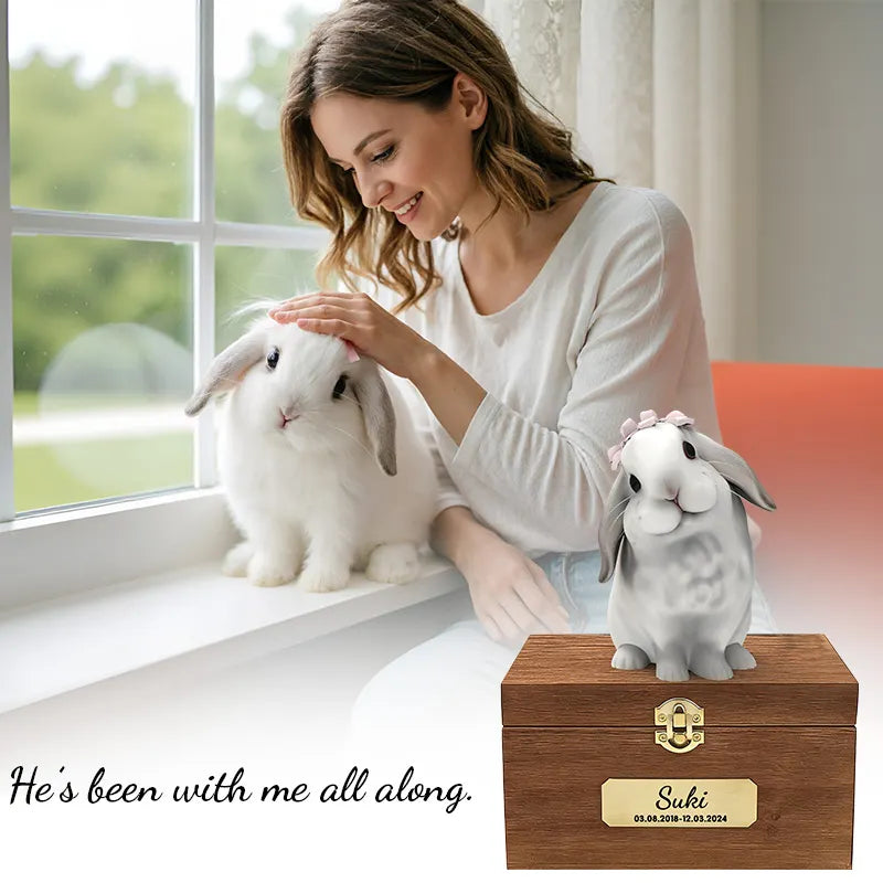 A woman smiles and pets a white lop-eared rabbit by a window. In front, the Bunny Sweet Memories wooden box holds a personalized pet urn with a plaque reading “Suki 01.09.2018–10.13.2024” and the words "He's been with me all along.